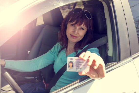 A smiling lady behind the wheel of her car shows her driver's license