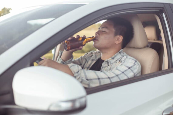 Young man drinking a bottle of beer behind the wheel of a car - cheap SR-22 insurance.